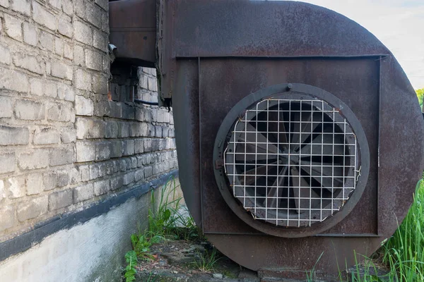 Industrial centrifugal radial exhaust fan of snail type of brown color with traces of rust, installed near building made of white silicate bricks with traces of soot, at side of which grass grows