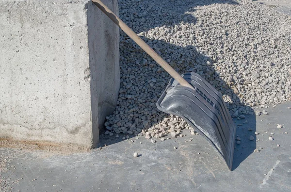 Tahta saplı geniş bir kürek ve çalışan siyah plastik bir parça gri çakıl yığınının yakınındaki düz bir yüzeyde beton bloğa yaslanıyor.