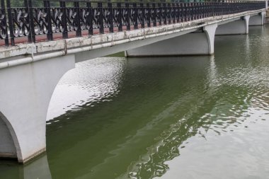 Küçük kemerli bir köprü perspektife girer. Yuvarlak şekilli beton sütunlar beyaza boyanır. Çit siyah metal parçalarından yapılmış. Aşağıda küçük dalgalı turkuaz sularda yansıma var..