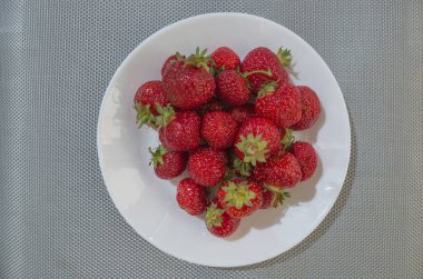 Bir demet olgun, olgun kırmızı-kestane çilekleri üst üste seramik bir tabağın içinde gri-gümüş bir hasır masa örtüsünün üzerinde duruyorlar..