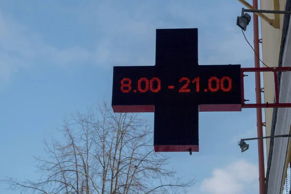 Eczanenin açık kırmızı haç işareti, LED lamba panelleri ile kutu şeklinde yapılmış, eczanenin açılış saatlerini gösteren, bulutlu kış gökyüzünün arka planındaki binanın ön cephesine kurulmuş