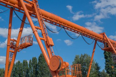 Orange metal single girder overhead crane with round beam cross-section, electrically driven, suspended cab with hook on two supports. In the background a row of trees and a blue cloudy sky.