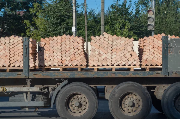 Bir kamyon, üzerinde kırmızı tuğlalar olan ahşap paletleri yol kenarındaki ağaçların arka planında trafik ışığı olan bir kavşaktan geçen uzun bir römorkun açık platformunda taşıyor..