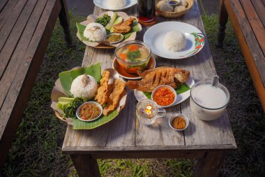 Endonezya yemekleri, sade ahşap bir masada servis edilir. Kızarmış balık, pilav, taze sebze, çorba ve açık hava yemek ortamında baharatlı sambal sunulur..