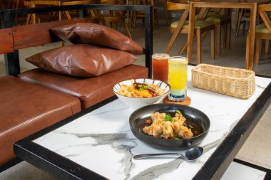 Samimi kafe masasında soslu tavuk katsu, çorbada hamur tatlısı, çıtır cips ve iki renkli buzlu içecek var. Deri koltuklu modern yemek seti.