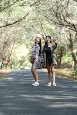 Two Asian Indonesian woman friends taking a selfie with a smartphone while standing on a quiet forest road, enjoying a cheerful outdoor moment surrounded by trees.