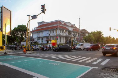 Gün batımında Yogyakarta, Endonezya 'da tarihi bir sömürge binasının önünden geçen arabalar ve motosikletlerle şehir kavşağı. Şehir trafiği, ulaşım, mimari ve günlük yaşamı gösteriyor..