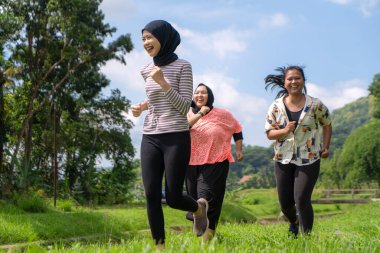 Üç neşeli Asyalı kadın ağaçlar, tepeler ve açık mavi gökyüzü ile çevrili çimenli bir arazide koşuyor..