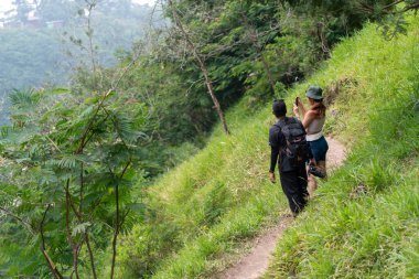 Endonezyalı çift verimli bir tropik orman patikasında yürüyüş yapıyor, doğanın tadını çıkarırken akıllı telefon kullanarak fotoğraf çekiyorlar. Açık hava macerası, seyahat yaşam tarzı ve eko turizm kavramı.