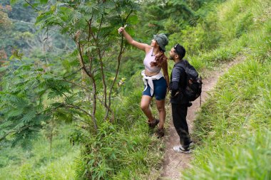 Endonezyalı çift, akıllı telefonlarıyla selfie çekerken verimli bir tropikal orman patikasında yürüyüş yapıyor. Açık hava macerası, seyahat yaşam tarzı ve doğa keşfi kavramı..