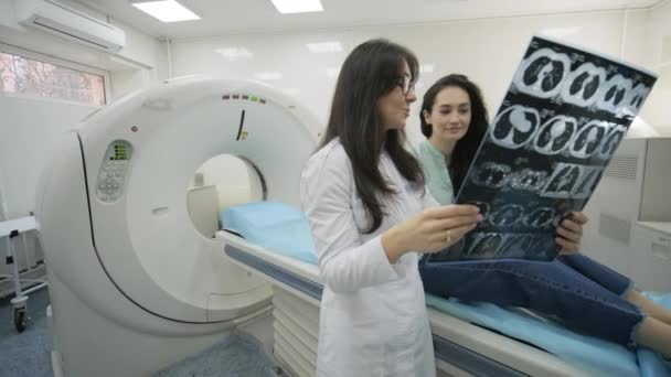 Vrouwelijke arts radioloog legt goede resultaten van CT-scan voor jonge vrouwelijke patiënt, toont de momentopname met beelden, observeren en analyseren van CT-scan in moderne kliniek naast moderne CT-scanner — Stockvideo