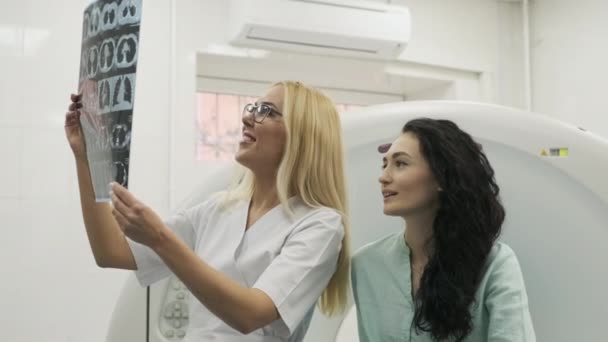 Kvinna läkare radiolog förklarar goda resultat av datortomografi för ung kvinnlig patient, visar ögonblicksbilden med bilder, observera och analysera datortomografi i modern klinik bredvid modern datortomografi — Stockvideo