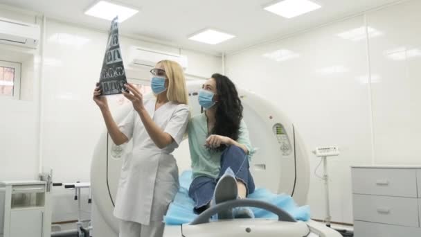 Lekarz radiolog wyjaśnia wyniki tomografii komputerowej w poszukiwaniu młodej pacjentki, prezentując migawkę z obrazami, obserwując i analizując tomografię komputerową w nowoczesnej klinice obok nowoczesnego skanera CT — Wideo stockowe