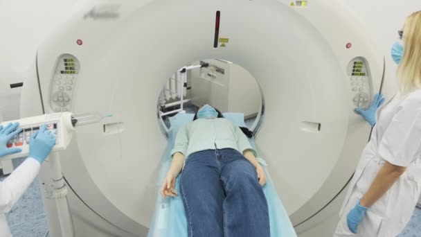 Kadın hasta, modern tıp kliniğinde maske ve eldiven takan iki kalifiye radyoloğun gözetiminde CT veya MRI taramasından geçiriliyor. Tomografide ya da MR masasında yatan hasta. — Stok video