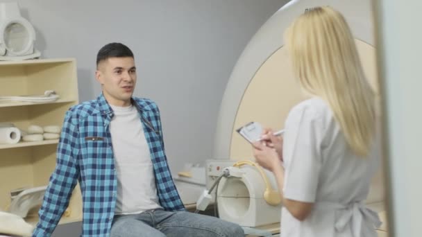 Kadın radyologlar genç erkek hastalarla diyalog kurar, hastanın tıbbi geçmişini sorar ve not alır, modern klinikte modern kapalı MR türünün yanında şikayetler — Stok video