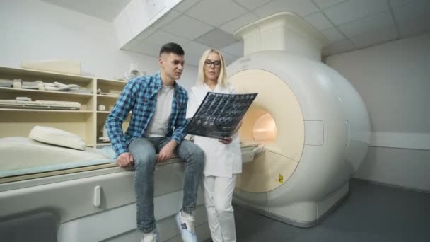Ärztin für Radiologie erklärt die Ergebnisse der MRT-Untersuchung bei jungen männlichen Patienten, zeigt den Schnappschuss mit Bildern, beobachtet und analysiert MRI-Scans in modernen Kliniken neben modernen geschlossenen MRT — Stockvideo