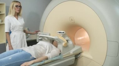 Kadın radyolog beyin, kafa ve boyun MR taraması yapıyor. Otomatik masadaki genç bayan hasta, modern ekipmanlarla kapalı bir MRI makinesine giriyor, hastanın kafasına dolanıyor..