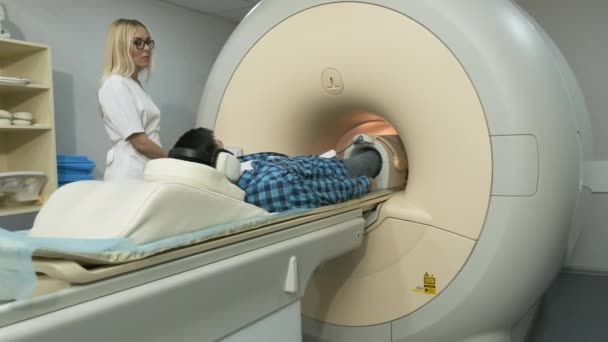 De vrouwelijke dokter maakt een knie-gewricht MRI scan. Jongeman patiënt op automatische tafel verlaat een gesloten MRI machine met behulp van geluidsisolatie koptelefoon. Moderne apparatuur, spoel op de knie van de patiënt — Stockvideo