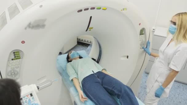 Paciente do sexo feminino é submetida a tomografia computadorizada ou ressonância magnética sob supervisão de dois radiologistas qualificados em máscara e luvas na clínica médica moderna. Paciente deitado em uma mesa de TC ou RM — Vídeo de Stock