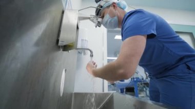 The surgeon prepares for the operation, washes and sterilizes his hands in the modern operating room. Doctor is equipped in mask, medical headlamp, surgical loupes