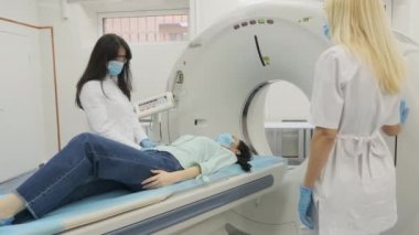 İki radyoloji uzmanı tomografi ya da MRI taraması yapılan kadın hastaya kontrast enjekte etmek için damar içi sonda yerleştiriyor. Hasta tomografide ya da MRI tarama yatağında yatıyor, makinenin içinde hareket ediyor..