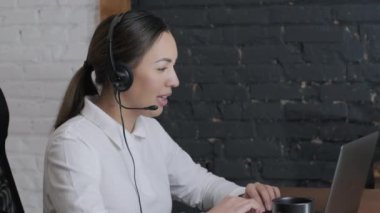 Gülümseyen kadın çağrı merkezi operatörü, müşteri destek ajanı kulaklık takıyor, danışmanlık yapıyor, dizüstü bilgisayarda yazıyor, internette konuşuyor, yardım hattı operatörü konferans videosu çekiyor.