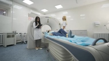 Kadın hasta, modern tıp kliniğinde iki kalifiye radyoloğun gözetiminde CT veya MRI taramasından geçiriliyor. Tomografide veya MR tarama masasında yatan hasta, makinenin dışına çıkıyor.