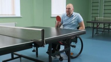 Tekerlekli sandalyedeki bir adam ping pong oynuyor. Özürlü insanlar masa tenisi oynar. Engellilerin rehabilitasyonu. Engelli sporu.