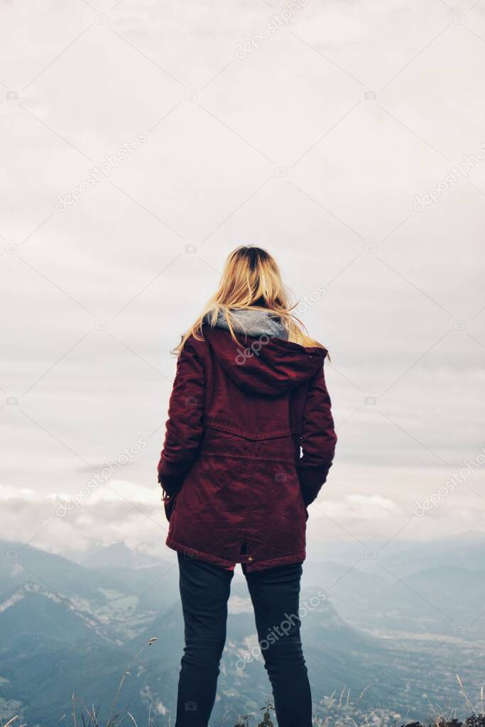 Young blond woman facing away standing on a cliff in winter wearing dark red burgundy coat looking into the distance to the foggy mountains