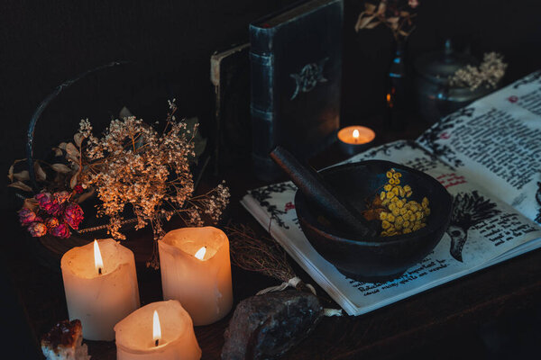 Wiccan witch altar prepared for casting a spell with an open Book of Shadows and pestle and mortar. Old spell books, crystals, burning lit candles, dried flowers in dark night background