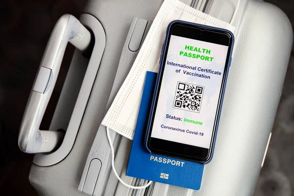 Smartphone with international vaccination certificate on screen, digital vaccination passport for COVID-19, qr-code, medical mask on suitcase. Health Passport concept, Coronavirus outbreak situation