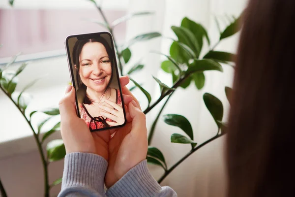 Smiling elderly mother and young daughter have video call, on mobile phone. Chat with happy mature mom, quarantine at home. Green leaves in background. Mother\'s day lockdown concept