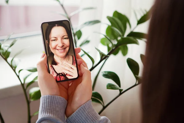 Smiling elderly mother and young daughter have video call, on mobile phone. Chat with happy mature mom, quarantine at home. Green leaves in background. Mother's day lockdown concept