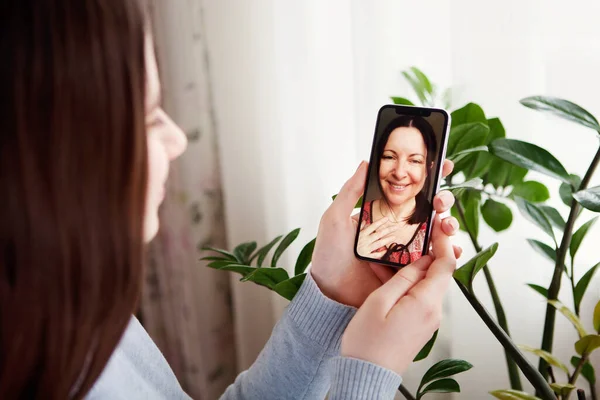 Gülümseyen yaşlı anne ve küçük kızı cep telefonundan görüntülü arama yapıyorlar. Mutlu olgun bir anneyle sohbet etmek, evde karantina. Arka planda yeşil yapraklar. Anneler Günü kilitleme kavramı