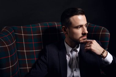 Düşünceli bir adamın portresi. Sakallı adam çenesine dokunuyor, sandalyede oturuyor. Siyah arkaplan.