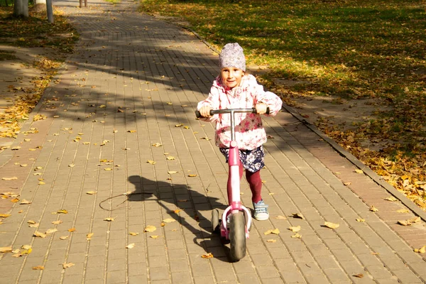 Ευτυχισμένο κορίτσι οδηγεί ένα σκούτερ σε ένα μονοπάτι στο πάρκο το φθινόπωρο — Φωτογραφία Αρχείου