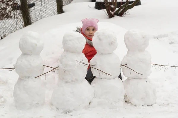 5年の少女が路上で冬の雪中に隠れています手の小枝を持った4人の雪だるま. — ストック写真