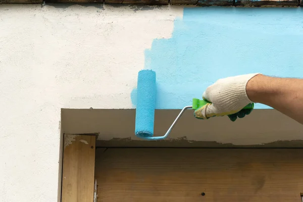 The man\'s hand paints a wall over a window or a door with blue paint