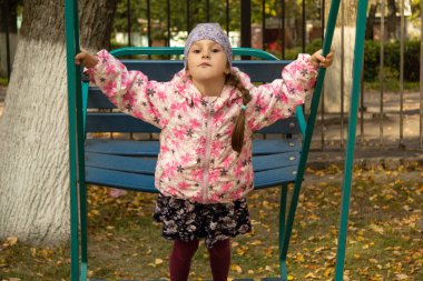 Beş yaşındaki kız, sonbaharda parkta büyük salıncaklarda sallanıyor.