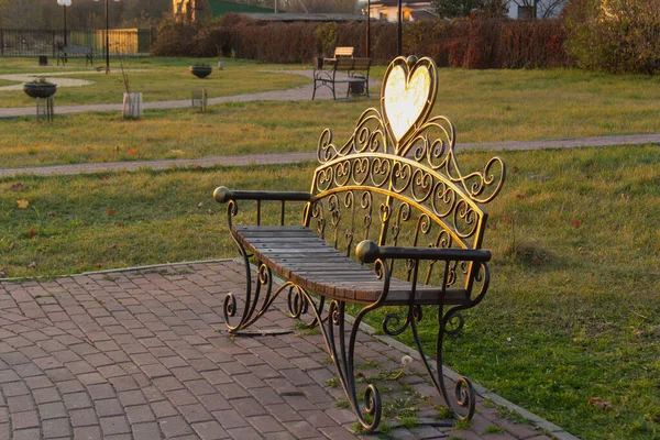 Panca concava in legno nel parco. Nei soli raggi il cuore di metallo brilla — Foto Stock
