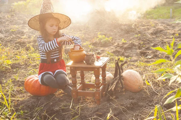 Uma menina em roupas de fada e um chapéu preto se misturam sobre um pote. Há fumo à volta dela. Halloween. — Fotografia de Stock