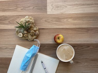 Not defteri, yüz maskesi, kahve, elma ve hava santrali olan ofis masaüstü çalışma alanı