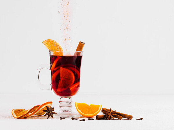 бокал горячего вина на светлом фоне со свежими и сушеными апельсинами, коричными палочками и наливкой порошка с корицей. Один из комплектов глинтвейна.