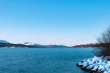 Kış gölü manzarası, suda dalgalanmalar, yerde kar, ufukta karla kaplı dağlar, ön plana odaklanma.