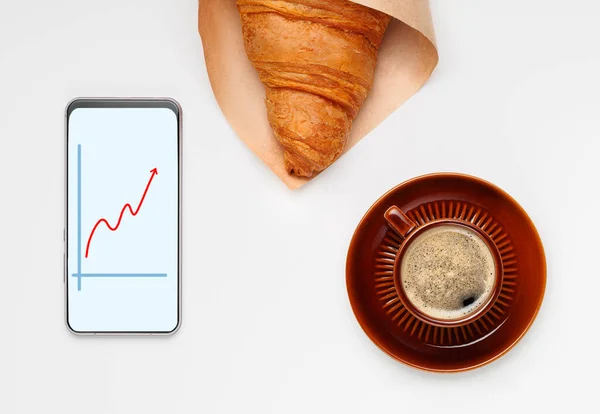 Bir fincan kruvasanla sade kahve ve borsa grafiğiyle telefon.