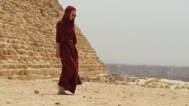 Uzun elbiseli bir kadın Giza piramidinin yanından geçiyor.