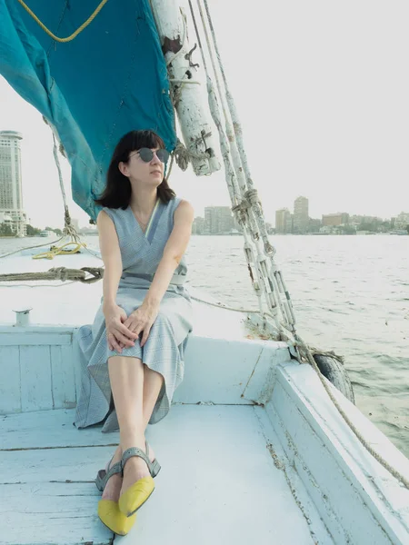 Mavi elbiseli ve güneş gözlüklü beyaz bir kadın Felucca teknesinde duruyor.