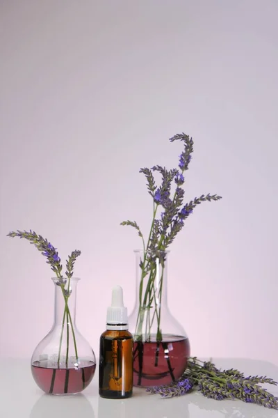 ラベンダーエッセンシャルオイルピペット付きのガラスボトル、紫色の背景にラベンダーの花と研究室のフラスコ。ラベンダーエキス配合の天然化粧品. — ストック写真