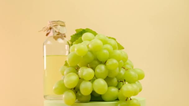 Олія виноградних кісточок. Скляна пляшка і купа зеленого винограду з листям на зеленому подіумі на бежевому фоні. Невелике обертання. Органічна натуральна біо виноградна олія — стокове відео