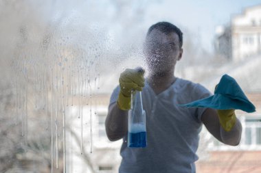 Elinde bez parçası olan tişörtlü bir adam cama deterjan sıkar. Camların pislikten temizlenmesi. Dezenfeksiyon ve hijyen.